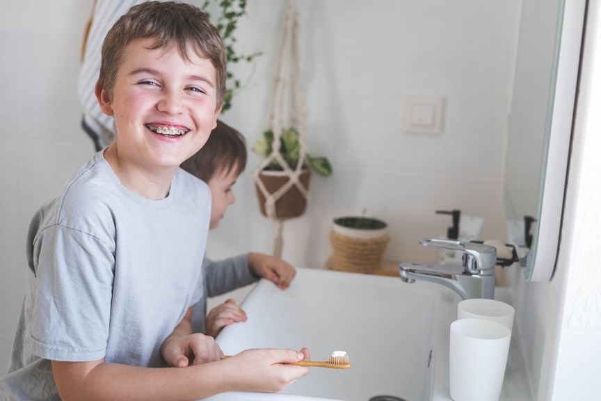 Lachendes Kind mit Zahnspange und Zahnbürste steht mit kleinem Bruder am Lavabo